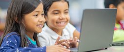 Two enthusiastic children are collaborating on laptops in a bright classroom setting, exhibiting teamwork and engagement while participating in a technology-based educational activity.